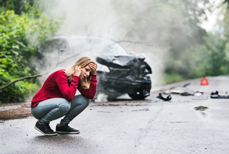 Woman in panic pose on phone reporting car crash on rural road with wrecked car in the background